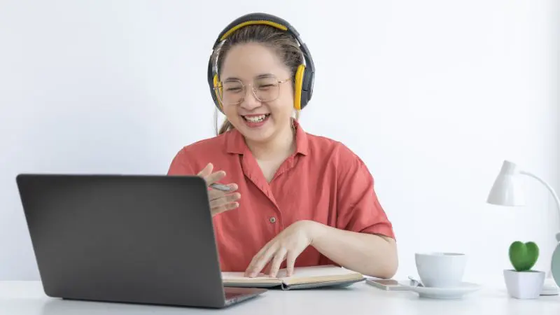 Smiling Asian girl in a orange t-shirt sitting at a desk while working on her laptop